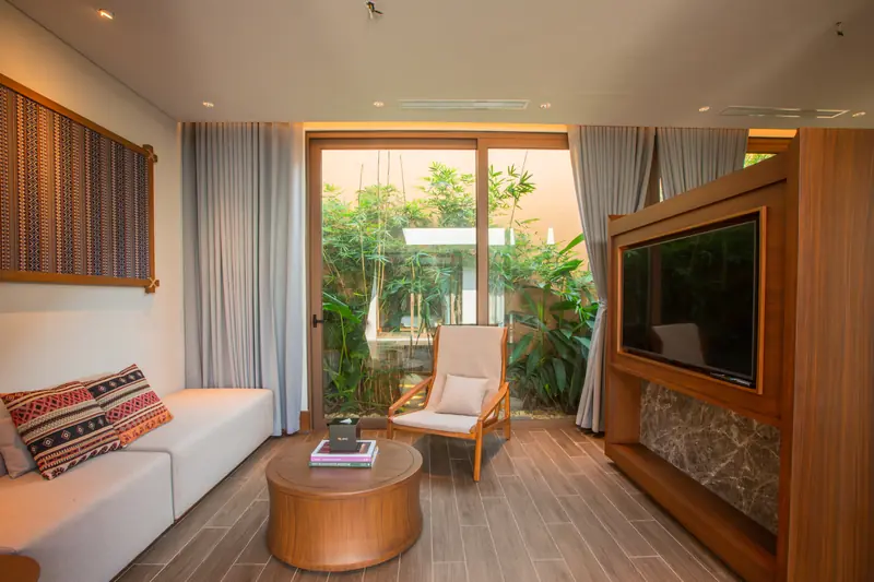 Phòng khách villa La Minh: sofa gối thổ cẩm, bàn tròn gỗ, tủ tivi, cửa kính nhìn ra vườn cây