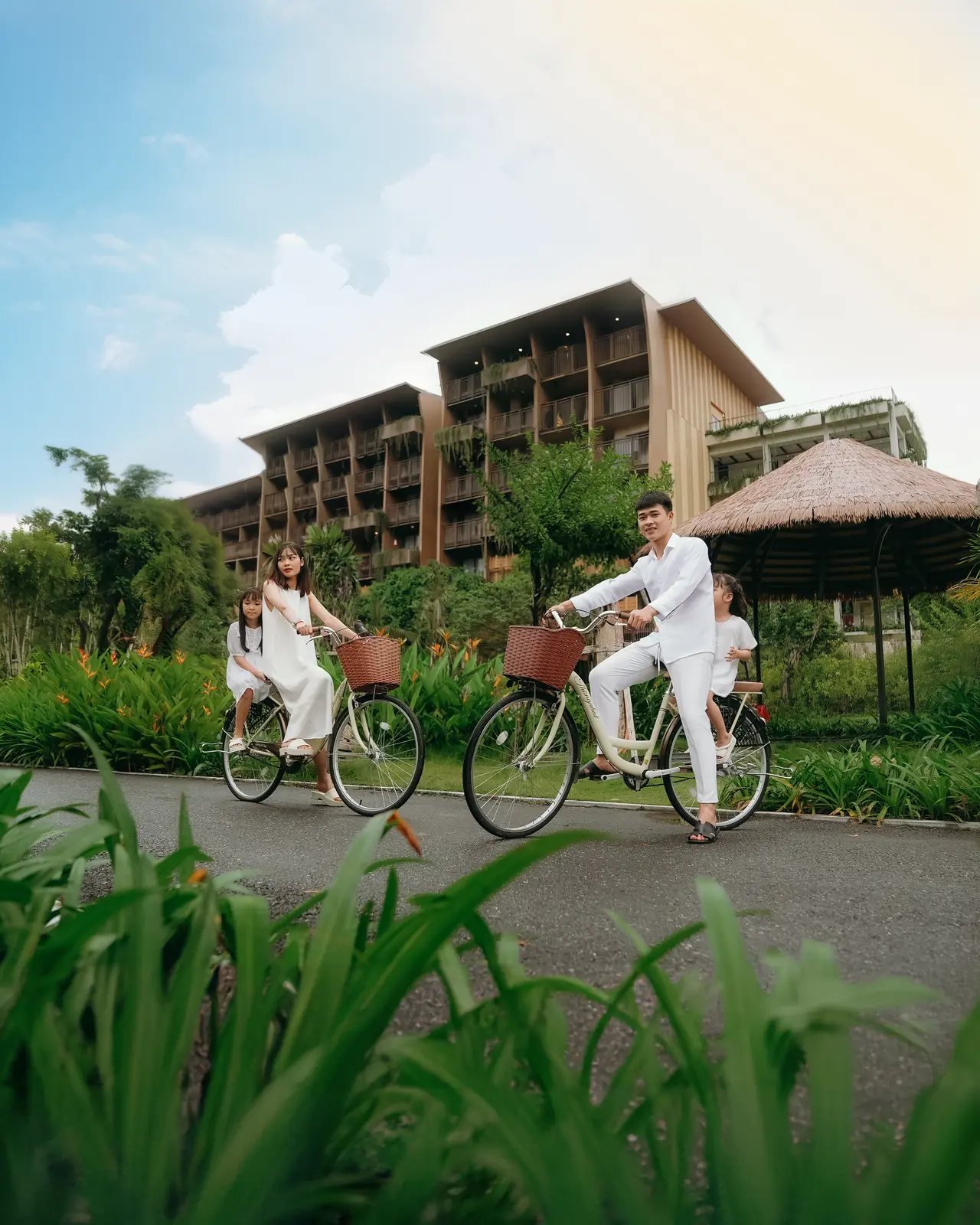 Gia đình đạp xe trên đường nội khu rợp cây, phía sau là tòa phòng nghỉ và chòi lá