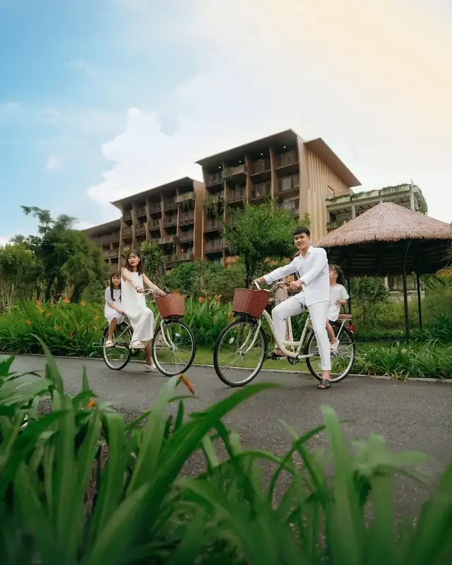 Gia đình bốn người đạp xe đạp có giỏ mây trên đường nội khu, hai bên là bụi hoa, phía sau là tòa phòng nghỉ và chòi mái lá