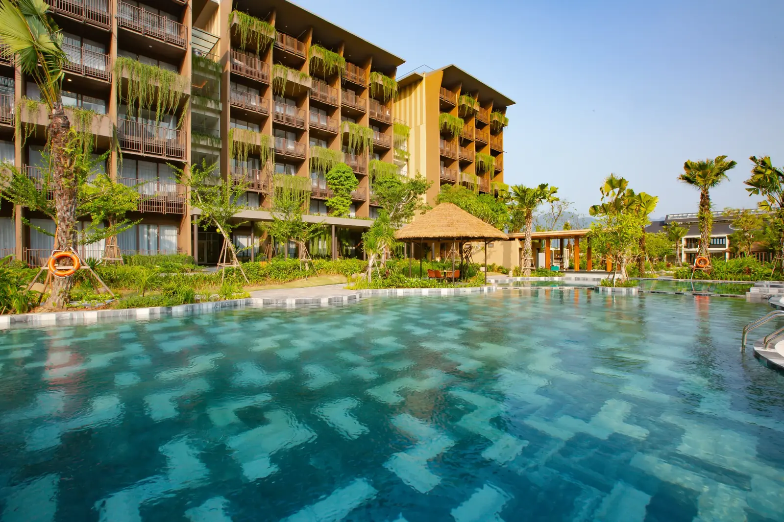 Mặt bể bơi lớn lát gạch xanh họa tiết, phía sau là tòa phòng phủ cây, hàng cọ và chòi lá