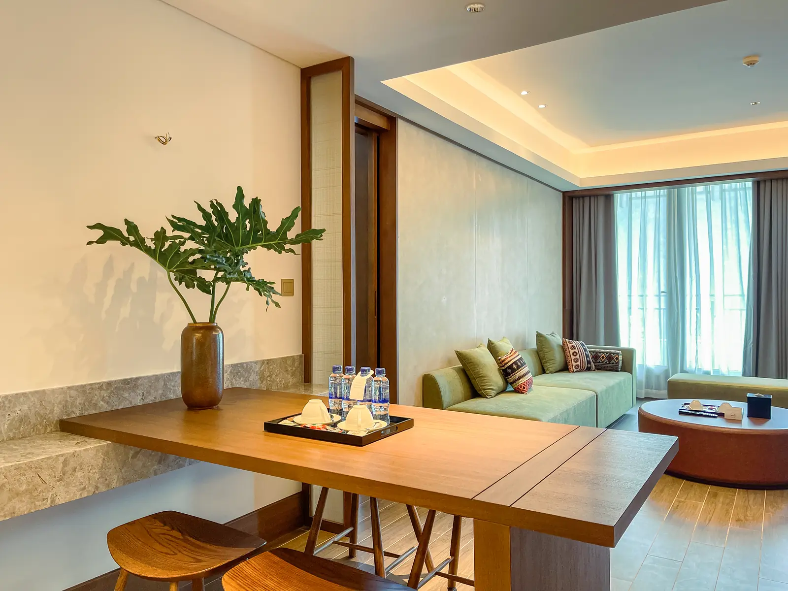 Bàn gỗ dài có ghế đôn trong phòng khách Family Suite, sofa xanh và cửa sổ rèm phía sau