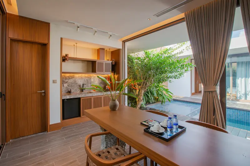Bàn ăn gỗ dài trong villa La Minh, cửa kính mở thẳng ra bể bơi riêng và cây xanh