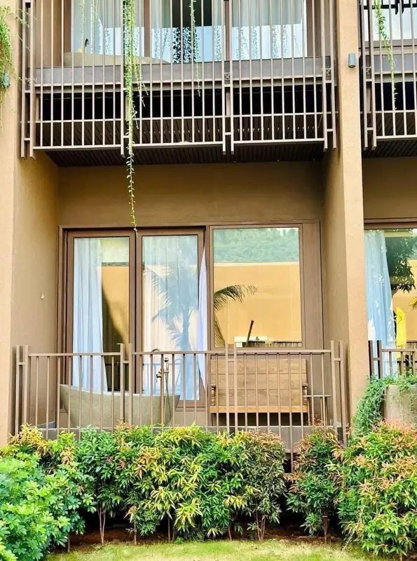 Mặt ngoài tòa phòng nghỉ: ban công tầng trệt có bồn tắm đá và ghế gỗ, hàng cây xanh trước lan can