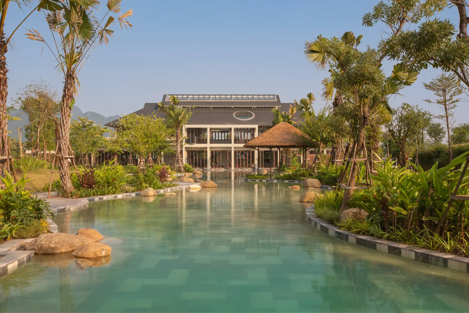 Hồ nước xanh giữa vườn nhiệt đới trước nhà đón tiếp, phía sau là núi