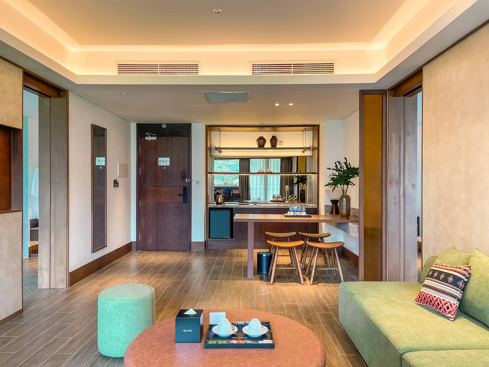 Phòng khách Family Suite: sofa xanh gối thổ cẩm, bàn tròn hồng đất, quầy bar nhỏ và ghế cao