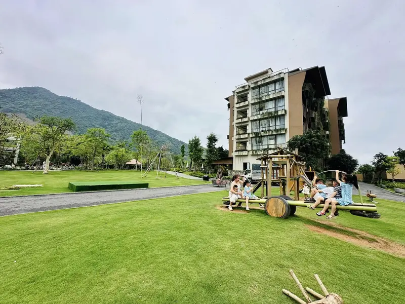 Sân cỏ rộng có đồ chơi gỗ, phía sau là tòa phòng nghỉ và núi