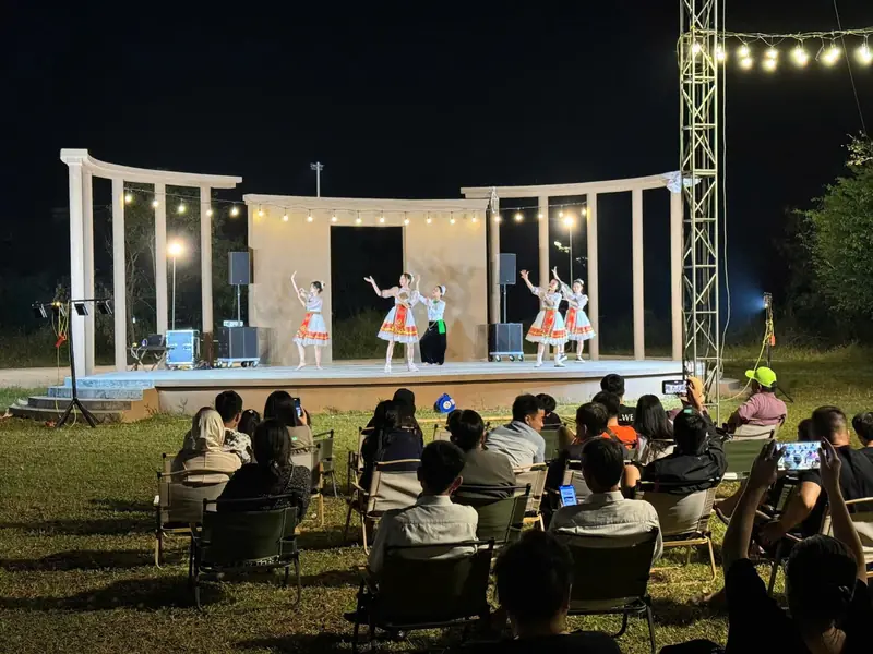 Sân khấu ngoài trời cột trắng sáng đèn dây, vũ công áo Mường biểu diễn, khán giả ngồi ghế dã ngoại