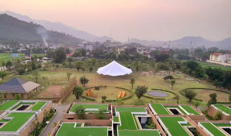 Nhìn từ trên cao xuống dãy villa mái phủ cỏ và bãi cỏ có lều trắng lúc chiều muộn, xa xa là núi